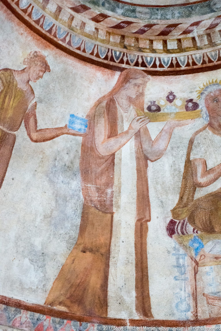 Painted standing attendant in a reddish tunic holding colorful vessels or offerings toward partially visible seated figures.