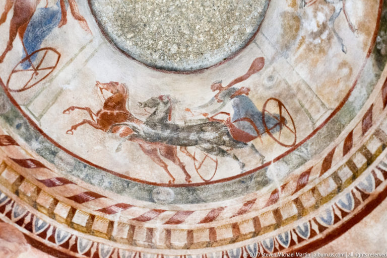Close upward view of a horse-drawn chariot with red wheel and blue-clad driver above patterned concentric borders.