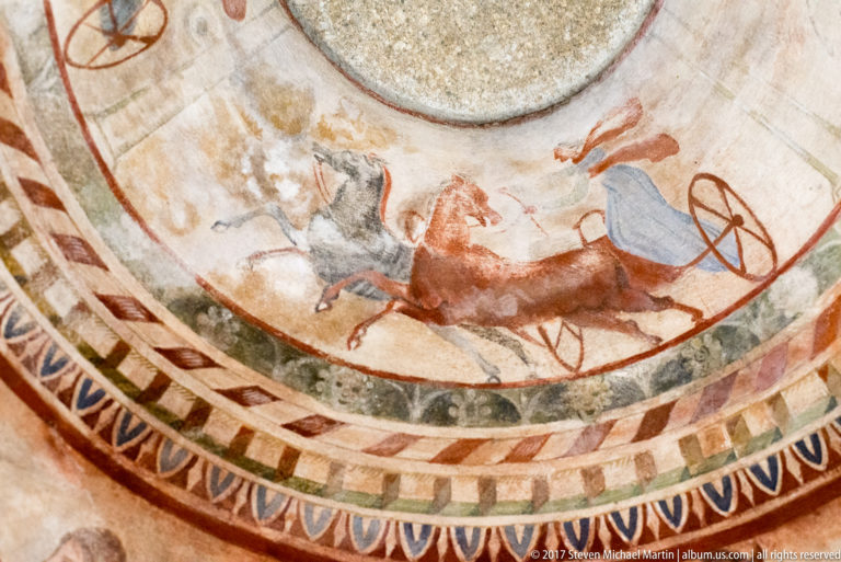 Upward view of a painted dome with horse-drawn chariot scene above concentric geometric borders in red, blue, black, and cream.