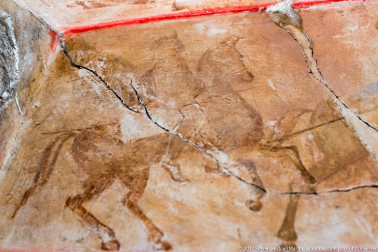 Faded wall painting of a horse and rider beneath a red border, crossed by large cracks and areas of plaster loss.