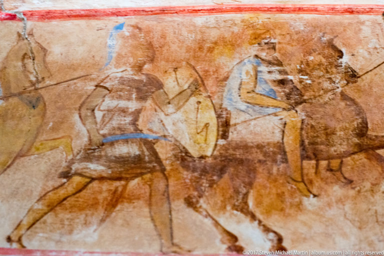 Painted horse in motion with a blue-and-ochre human figure above, crossed by cracks and worn areas in the plaster.