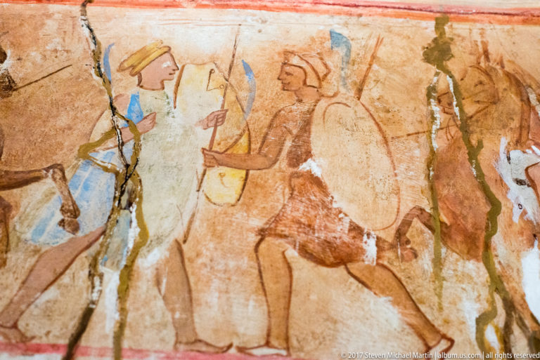 Close view of painted processional figures in profile wearing blue, ochre, red, and cream garments on cracked plaster.