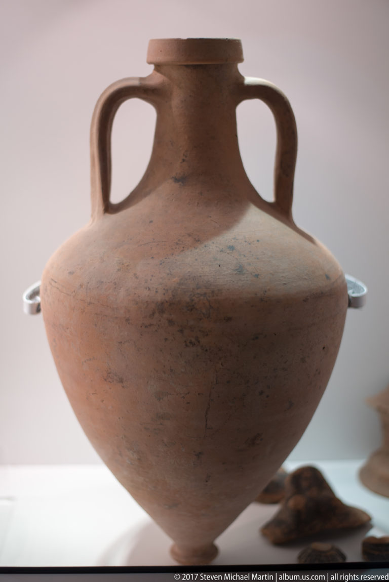 Large orange terracotta vessel with broad pointed body, narrow neck, flat rim, and two tall loop handles.