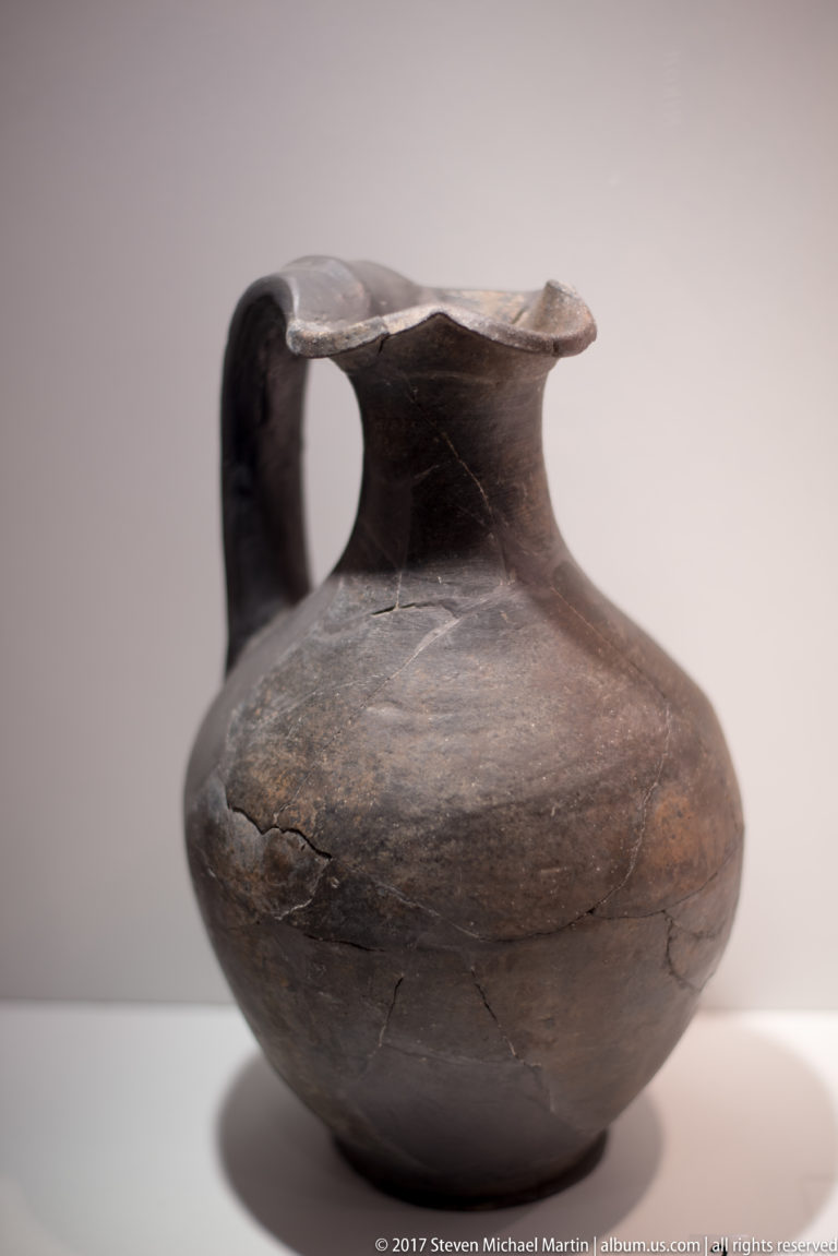 Squat dark brown ceramic jug with one handle, short neck, flared rim, rounded body, and visible surface cracks.