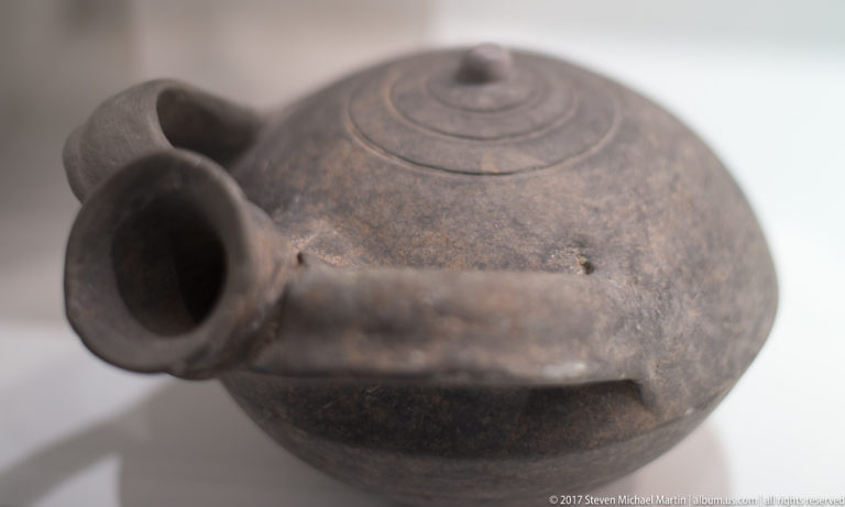 Dark round-bodied ceramic jug with loop handle, tubular projection, and concentric incised circles on the upper surface.