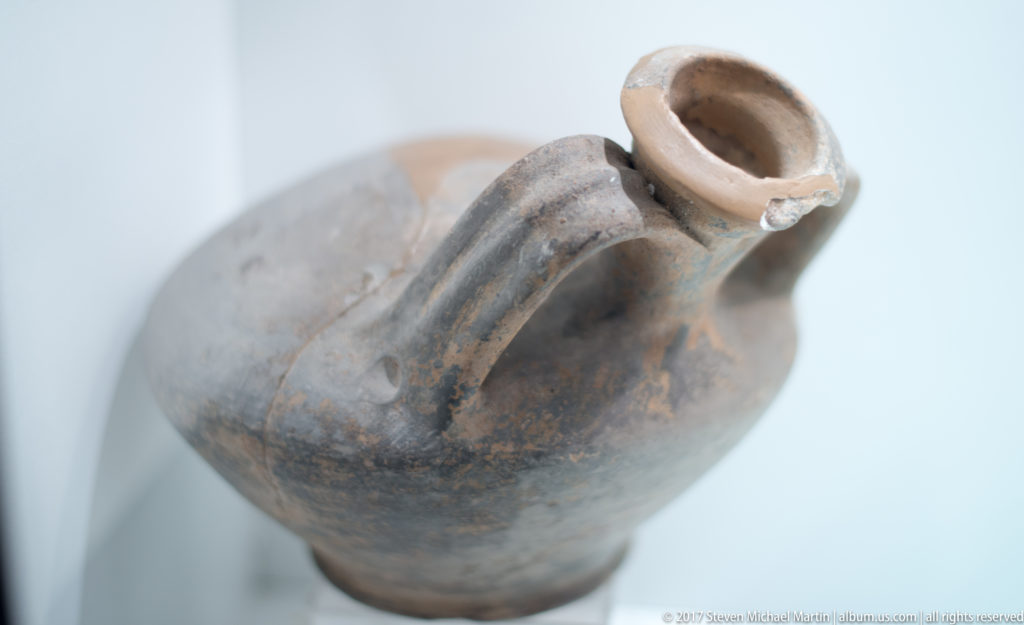 Oblique view of a small gray-black ceramic vessel with narrow neck, handle, raised longitudinal ribs, and a chipped rim.