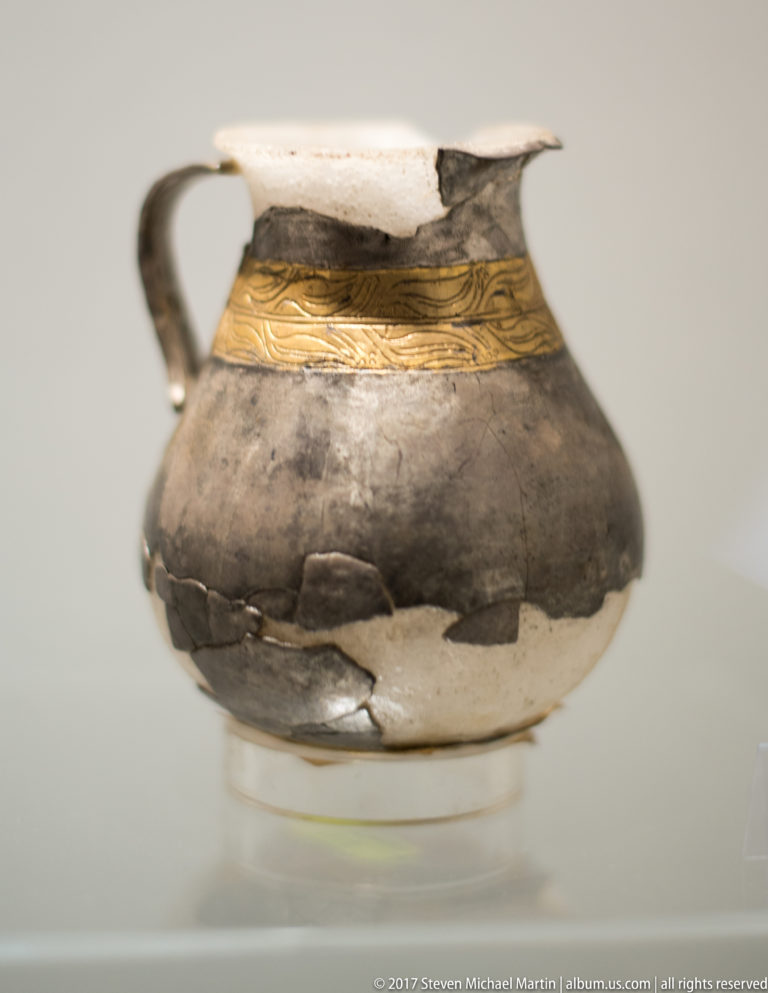 Small dark ceramic jug with one handle, repaired cracks, pale losses, and a narrow gold-decorated band around the shoulder.