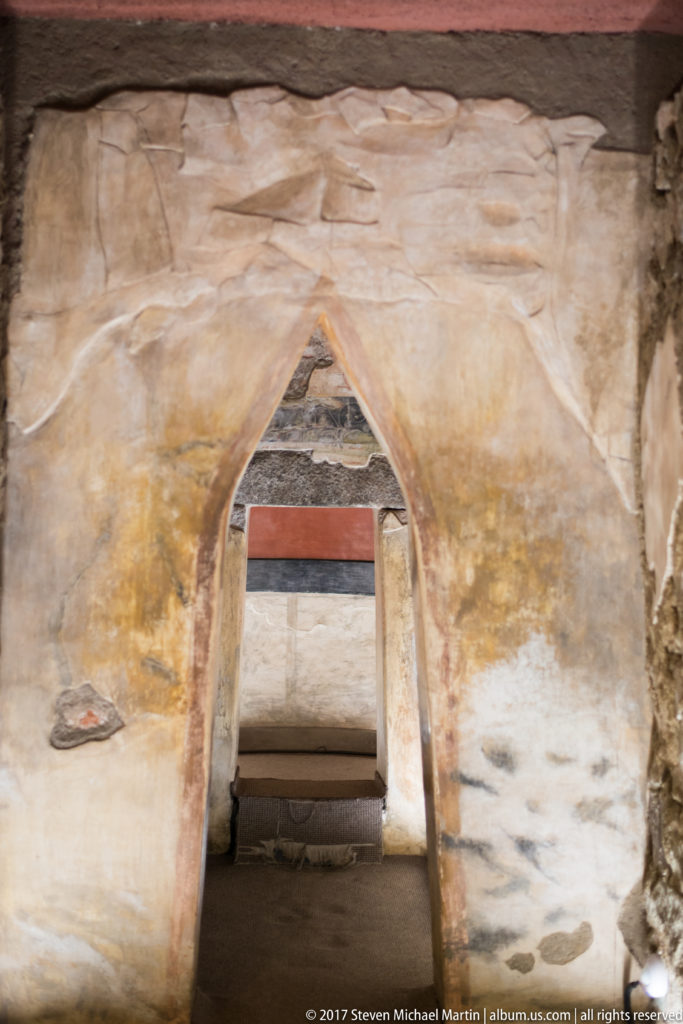 Narrow doorway inside a painted tomb interior with ochre plaster, red and dark horizontal bands, and a low passage beyond.