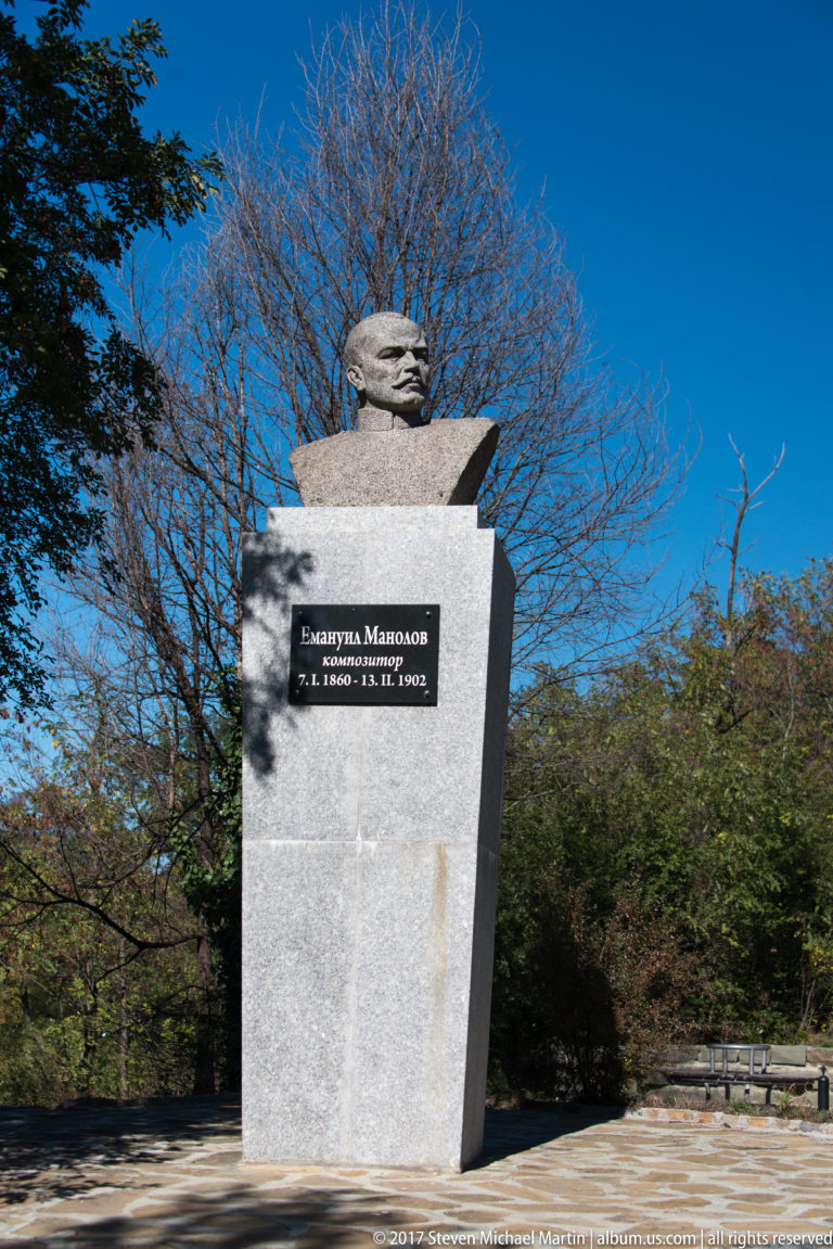 Bust of composer Emanuil Manolov on a tall stone pedestal with a black Cyrillic inscription plaque.