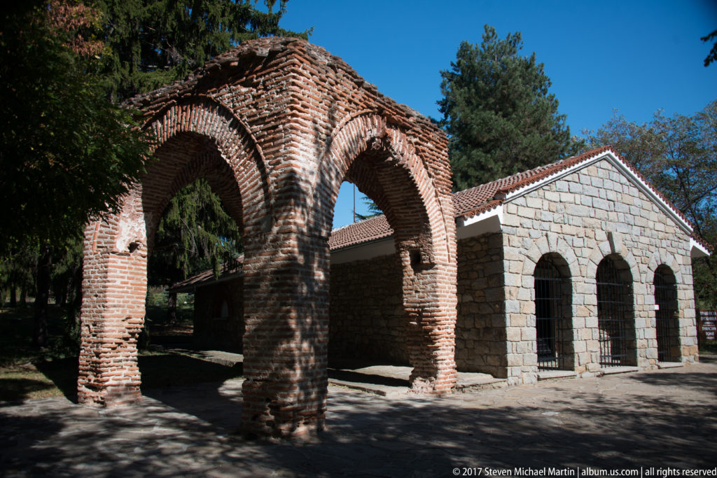 Oblique view of a brick arched gateway beside a gabled stone building with barred openings at the Kazanlak tomb site.