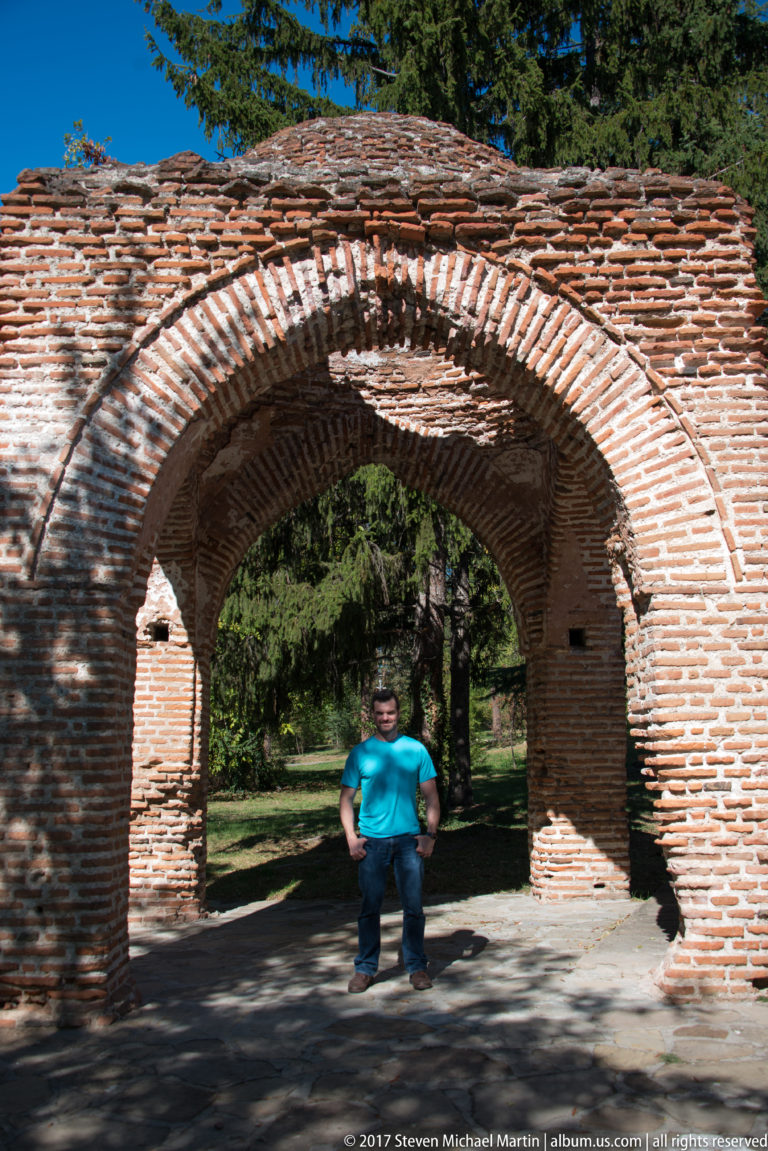 Large brick archway with a visitor standing beneath it, opening onto trees and lawn at the Kazanlak tomb site.