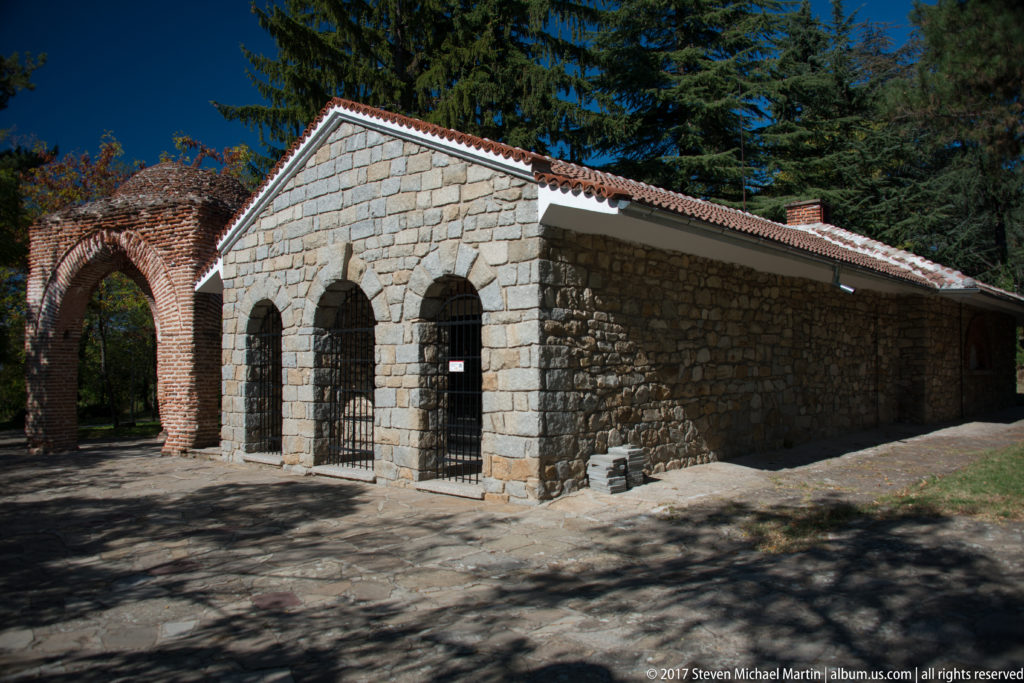 Stone gabled building with barred arched openings beside a brick gateway on the grounds of the Thracian Tomb of Kazanlak.
