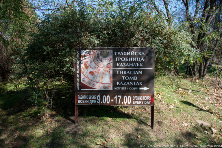 Bilingual Thracian Tomb of Kazanlak sign among trees, with opening hours, painted-tomb graphic, and arrow pointing right.