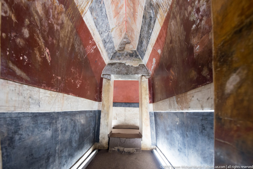 Centered view down a narrow painted corridor with dark lower walls, white and red bands, and a low doorway ahead.