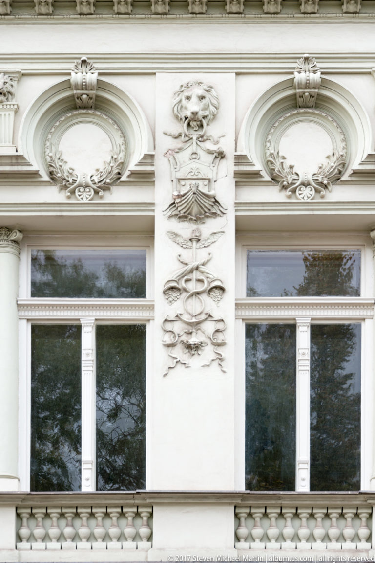 Pale stucco relief with a stylized face and scrollwork between two ornate upper-story windows.