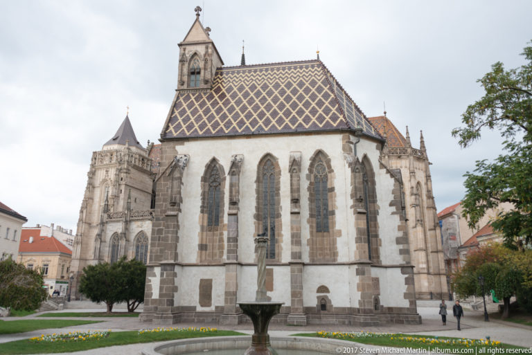 Broad side view of St. Elisabeth Cathedral with tall Gothic windows, patterned roof, masonry buttresses, and a low fountain.