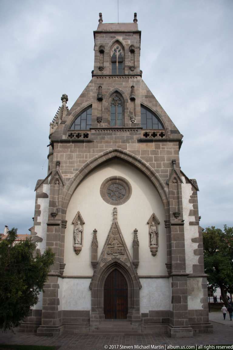 Frontal church façade with dark stone trim, pale stucco, pointed entrance, circular window, statues, and stepped upper gable.