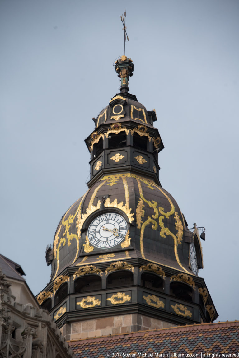 Black domed tower decorated with gold scrollwork and multiple clock faces above a red-tiled roofline.