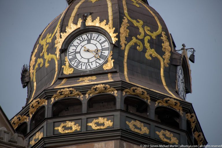 Close view of a clock face set into a black domed tower with elaborate gilded scrollwork and arched openings.