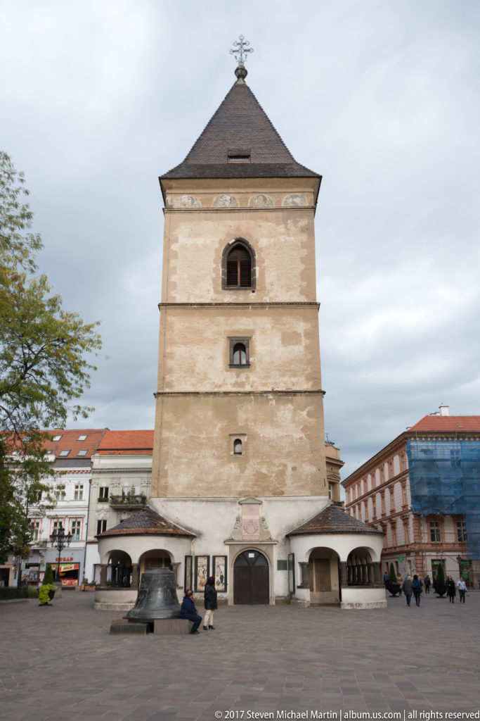 Freestanding square Urban Tower with pale weathered walls, pyramidal roof, small openings, and a large bell displayed at its base.