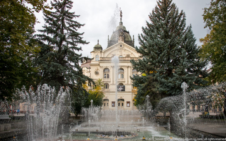 Singing Fountain spraying in front of the ornate State Theatre Košice, partly framed by tall evergreen trees.