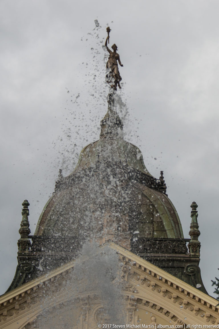 Rooftop statue and domed theater roof partly obscured by rising spray from the Singing Fountain.
