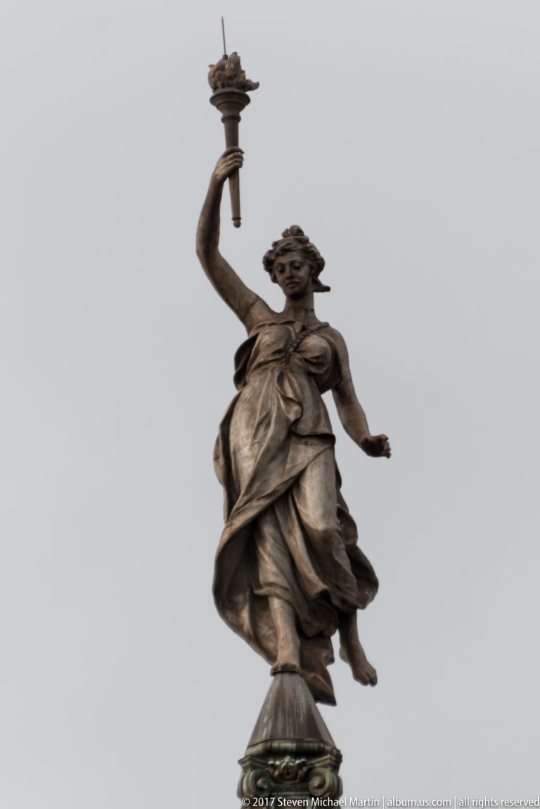 Dark rooftop statue of a draped female figure raising a torch-like object against a gray sky.
