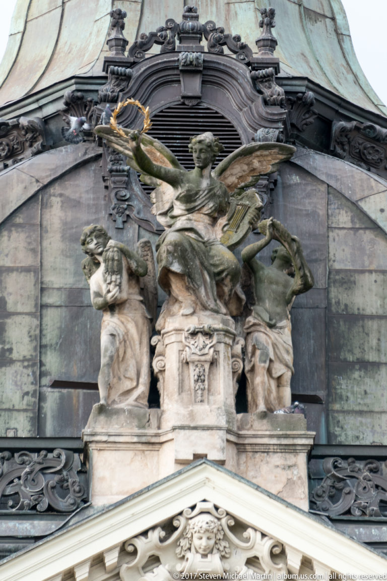 Winged rooftop figure above two seated sculptures amid dark metal roofing and ornate architectural details.