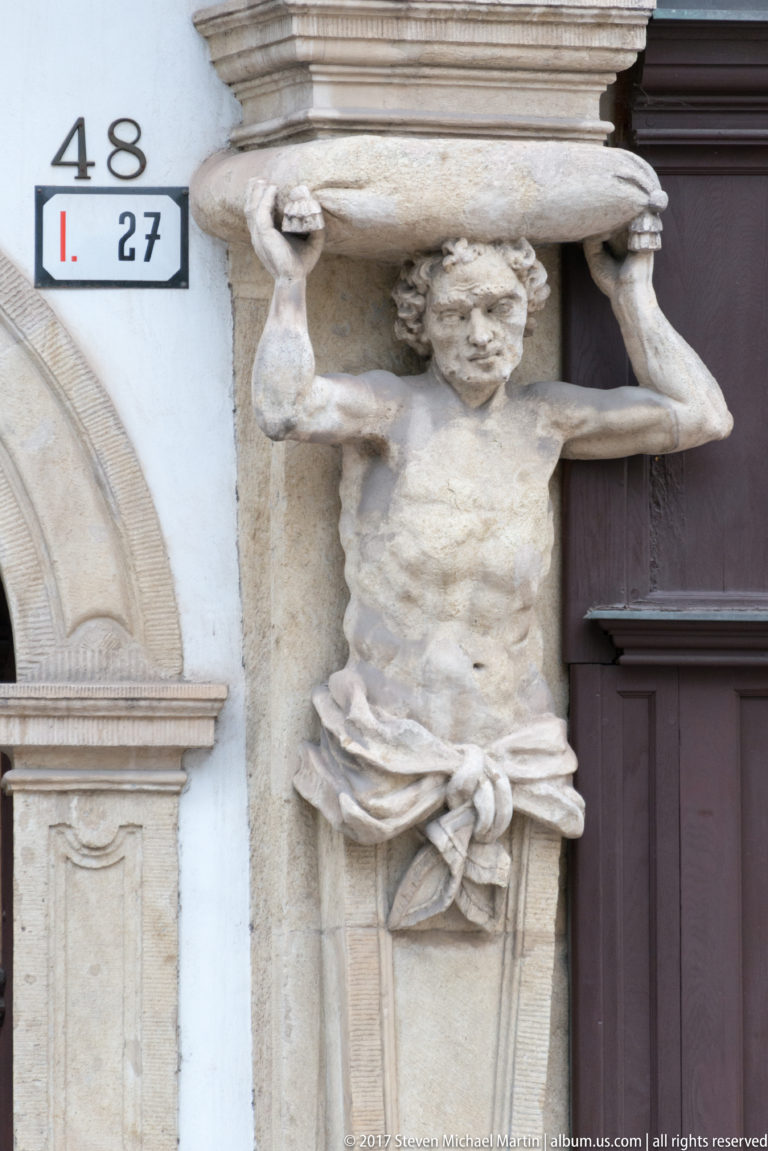 Carved male atlas figure beside a doorway, arms raised to support a projecting stone block above his head.