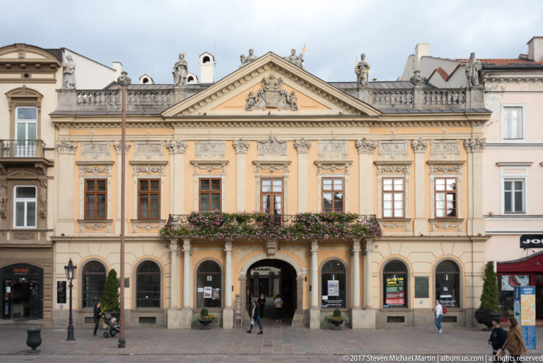 Symmetrical ochre historic building with arched ground floor, central passage, flowered balcony, tall windows, and sculpted pediment.