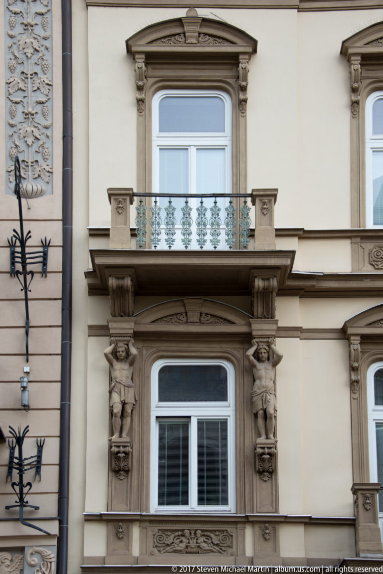 Two carved male atlas figures flank a window below a projecting balcony on a historic Košice façade.
