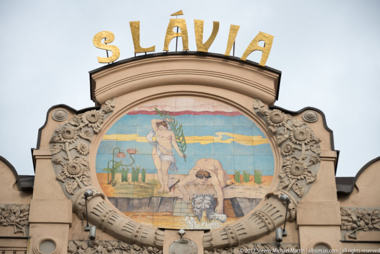 Gold SLÁVIA letters above a colorful figurative mosaic framed by dense sculpted ornament on a historic façade.