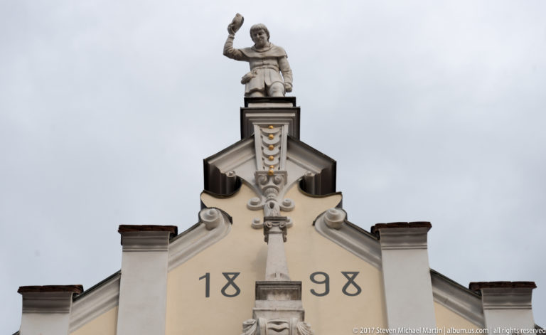Historic gable dated 1898, topped by a standing male statue with one arm raised against a cloudy sky.