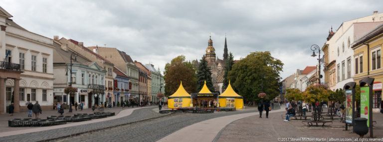 Curving pedestrian street lined with historic buildings and yellow tents, with cathedral towers above trees ahead.