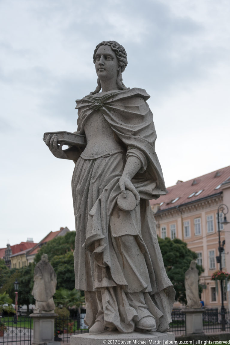 Stone draped female statue holding a circular object, with other sculptures and historic buildings blurred behind.
