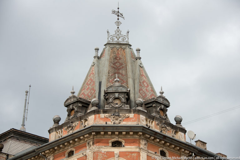 Ornate corner roof tower with patterned tiles, finials, sculptural trim, and metal cresting against a gray sky.