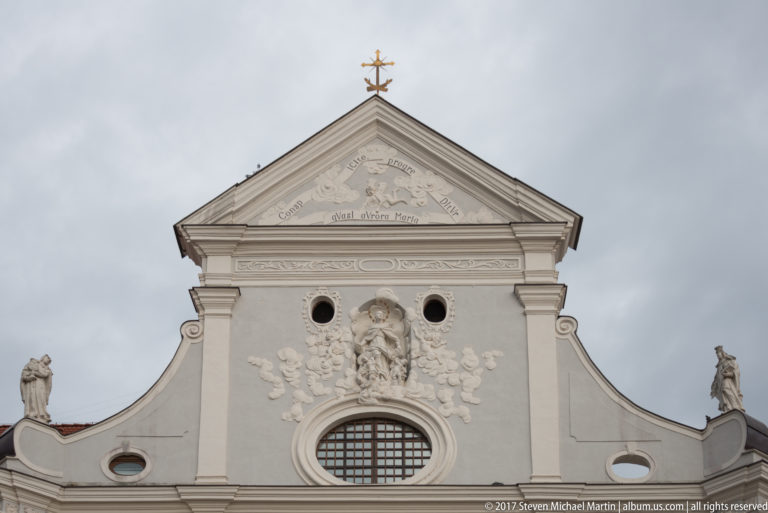 Upper Baroque church façade with oval window, sculptural relief, statues, triangular pediment, and cross.