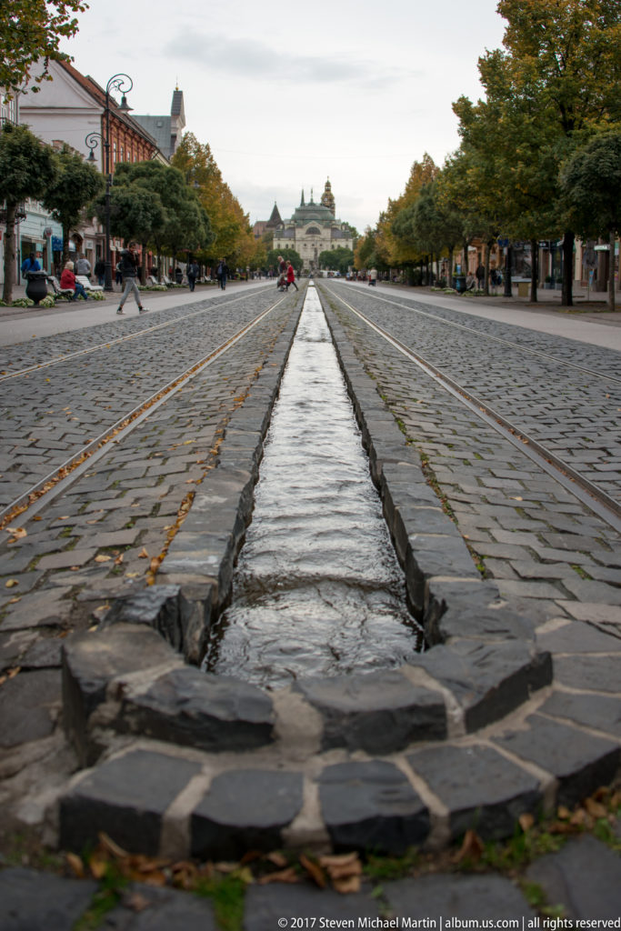 Stone-lined water channel receding through a broad cobbled promenade with rails, trees, pedestrians, and distant buildings.
