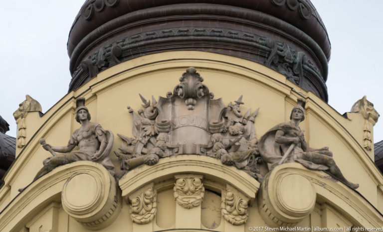Two seated stone figures flank an ornate cartouche beneath the dark base of a domed roof.