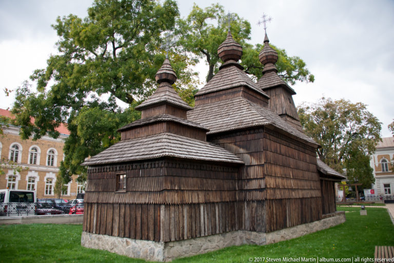 Traditional wooden church with shingled walls, tiered roof towers, and crosses standing on a grassy site.