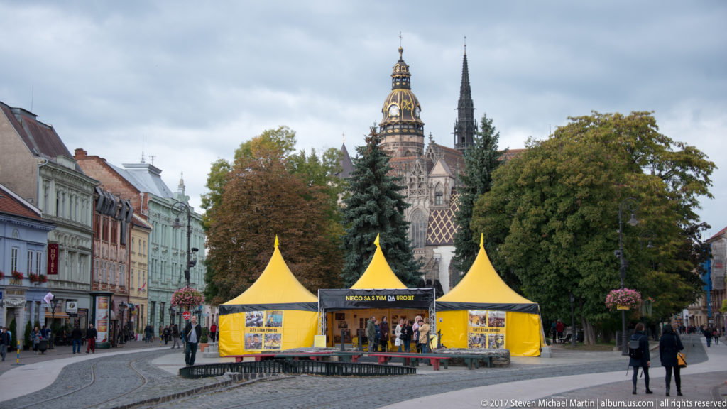 St. Elisabeth Cathedral towers and patterned roof rising behind trees and yellow market tents on Hlavná ulica.