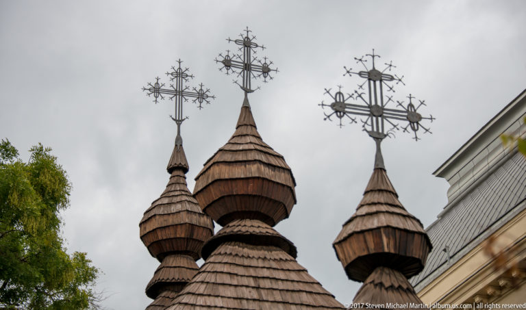 Three shingled wooden church towers topped by elaborate openwork metal crosses against a cloudy sky.
