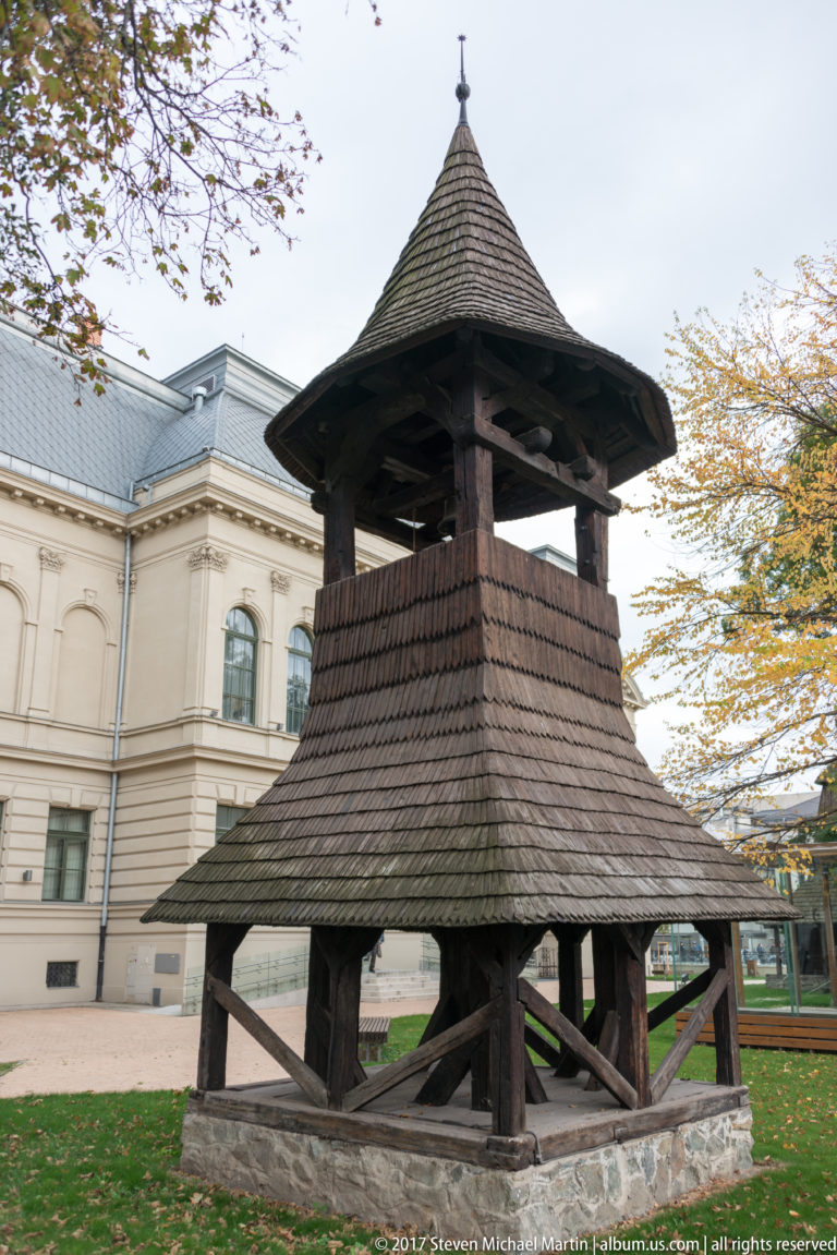 Open timber bell tower on a stone base with heavy bracing and a broad pyramidal shingled roof.