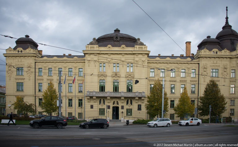 Broad symmetrical historic civic building with central entrance, balcony, flags, and dark roof pavilions along a city street.