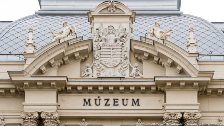 Ornate East Slovak Museum pediment with MÚZEUM inscription, central coat of arms, sculpted figures, and roof cresting.