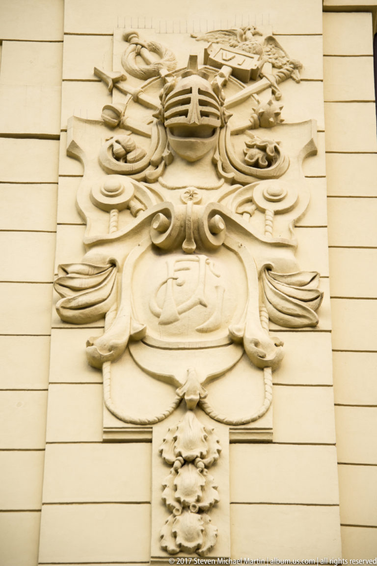Large pale heraldic relief with shield, helmet, crest, flowing mantling, and floral pendant on a façade.