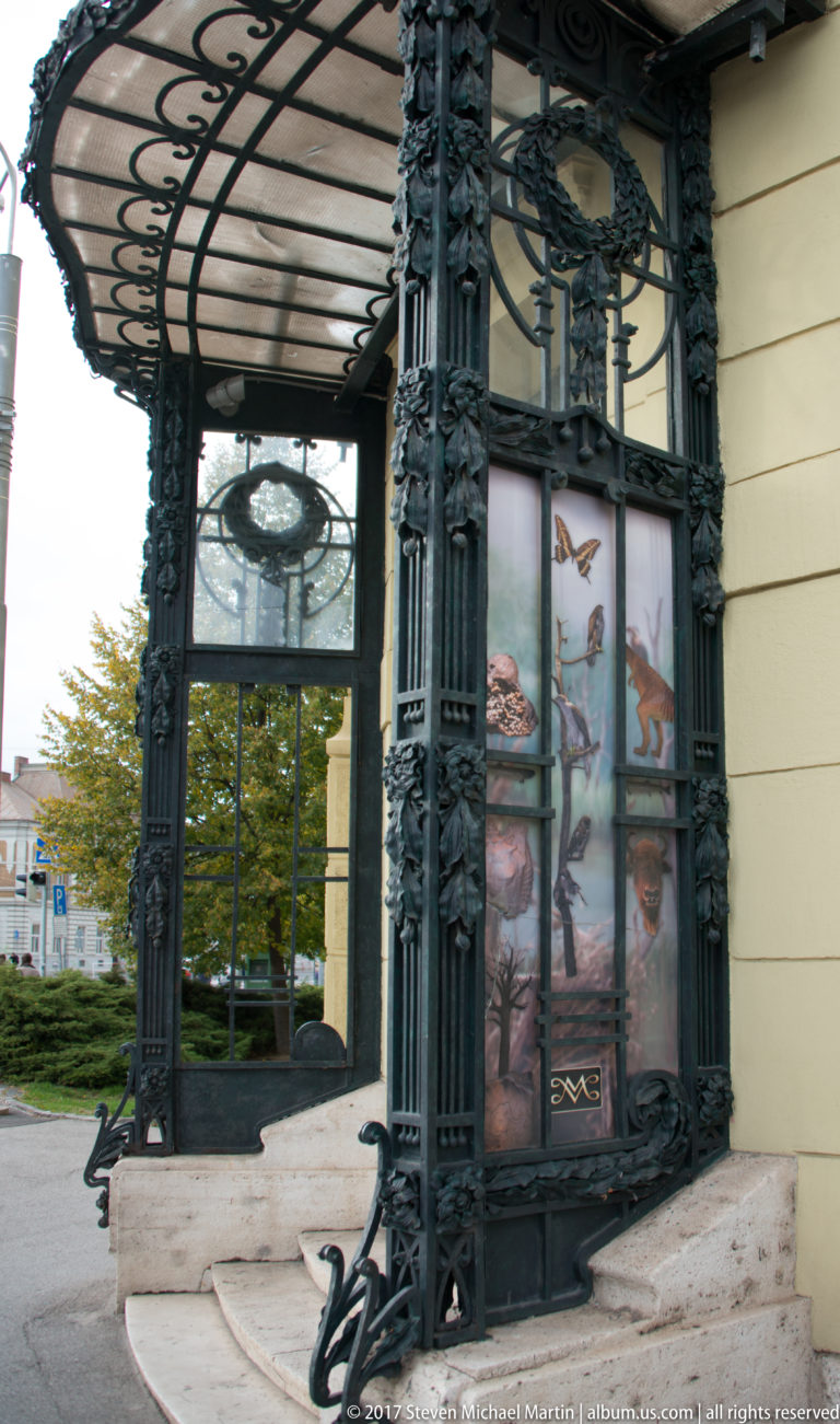 Ornate dark cast-iron entrance canopy with glazed panels, decorative columns, curved brackets, and stone steps.