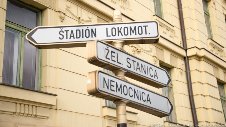 White arrow signs reading ŠTADIÓN LOKOMOT., ŽEL. STANICA, and NEMOCNICA mounted against a pale building.