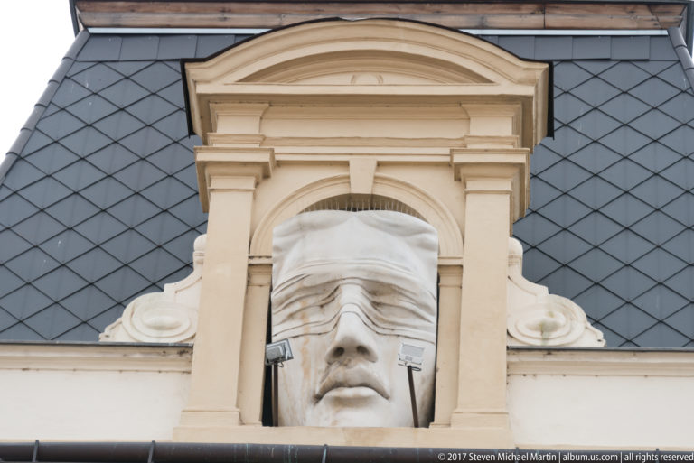 Large sculpted face with a band covering its eyes, set in a pale architectural niche beneath a curved pediment.