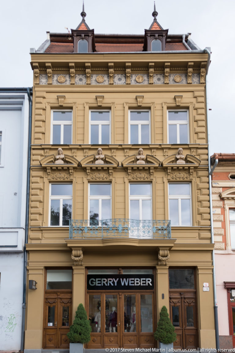 Ochre historic façade with carved window surrounds, pilasters, cornices, wrought-metal balcony, and ground-floor storefront.